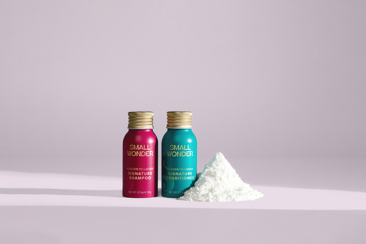 Two small, vibrant teal and magenta bottles of Small Wonder Signature Shampoo and Conditioner stand next to a neat mound of white concentrated powder on a light pink surface